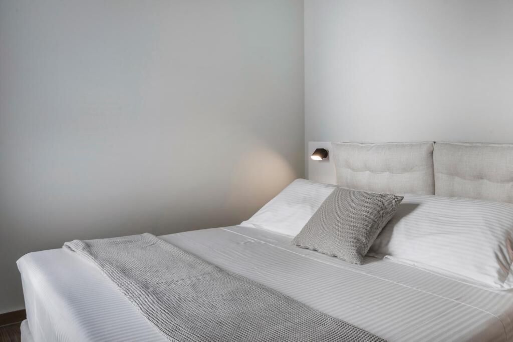 Una cama bien tendida con sábanas blancas y una manta gris, un aplique de pared y una pared lisa.