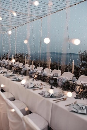 A long table set for a meal with chairs, under a canopy with string lights, and a view of the outdoors.