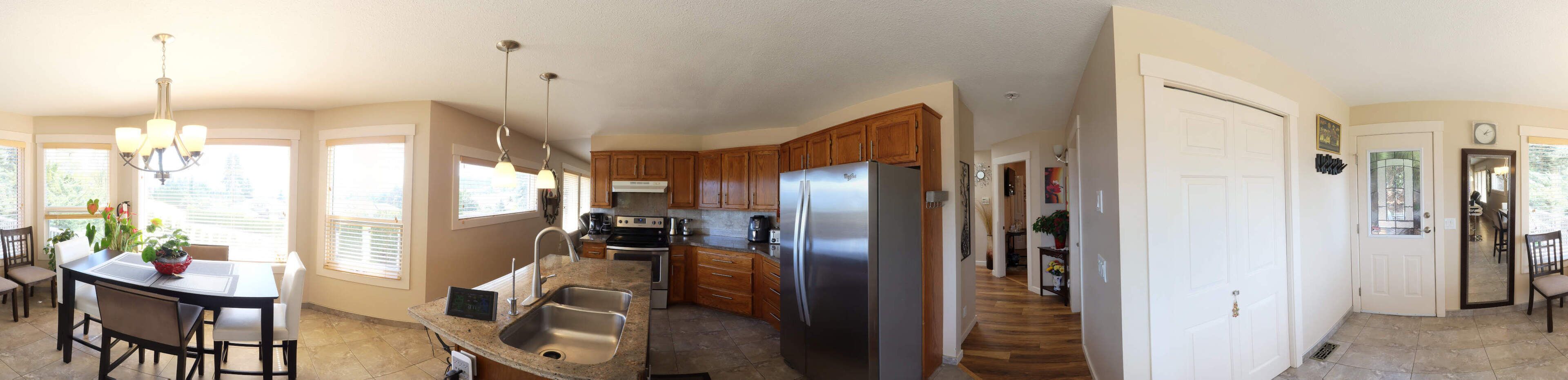 Una cocina con armarios de madera, electrodomésticos de acero inoxidable, una zona de comedor con mesa y sillas, y un pasillo con una puerta y un reloj.