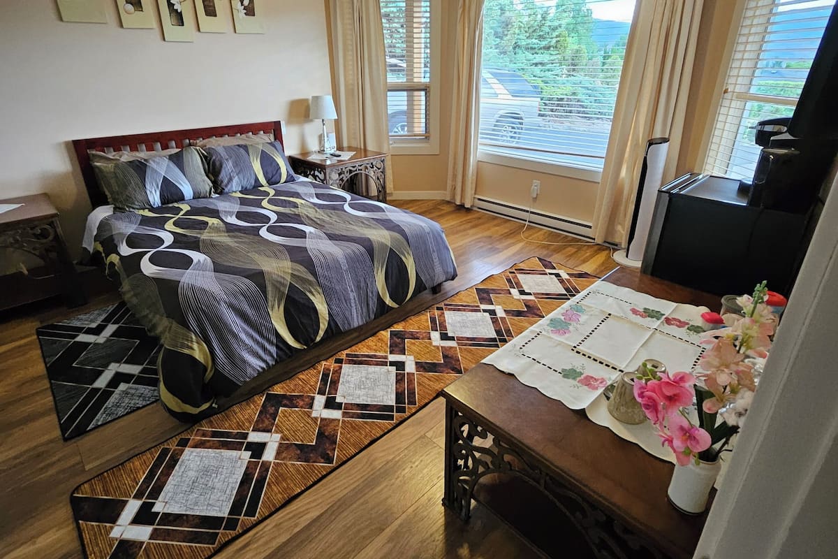Une chambre à coucher avec un lit, une table sur laquelle est posé un vase, une fenêtre avec des rideaux et une vue sur l’extérieur.