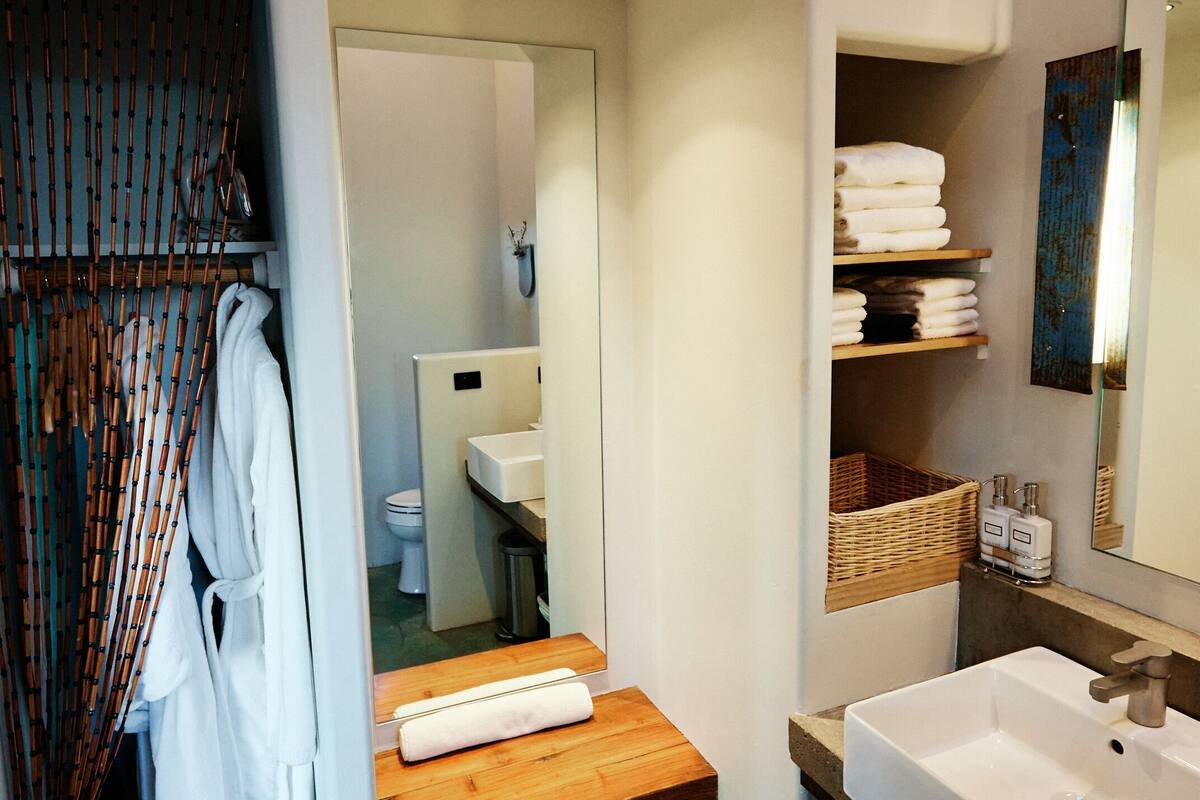 Une salle de bain avec une étagère en bois, un lavabo blanc, un miroir et des toilettes visibles à travers une porte ouverte.