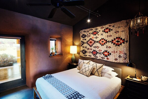 A bedroom with a bed, a window, a ceiling fan, a wall hanging, and a nightstand with a lamp.