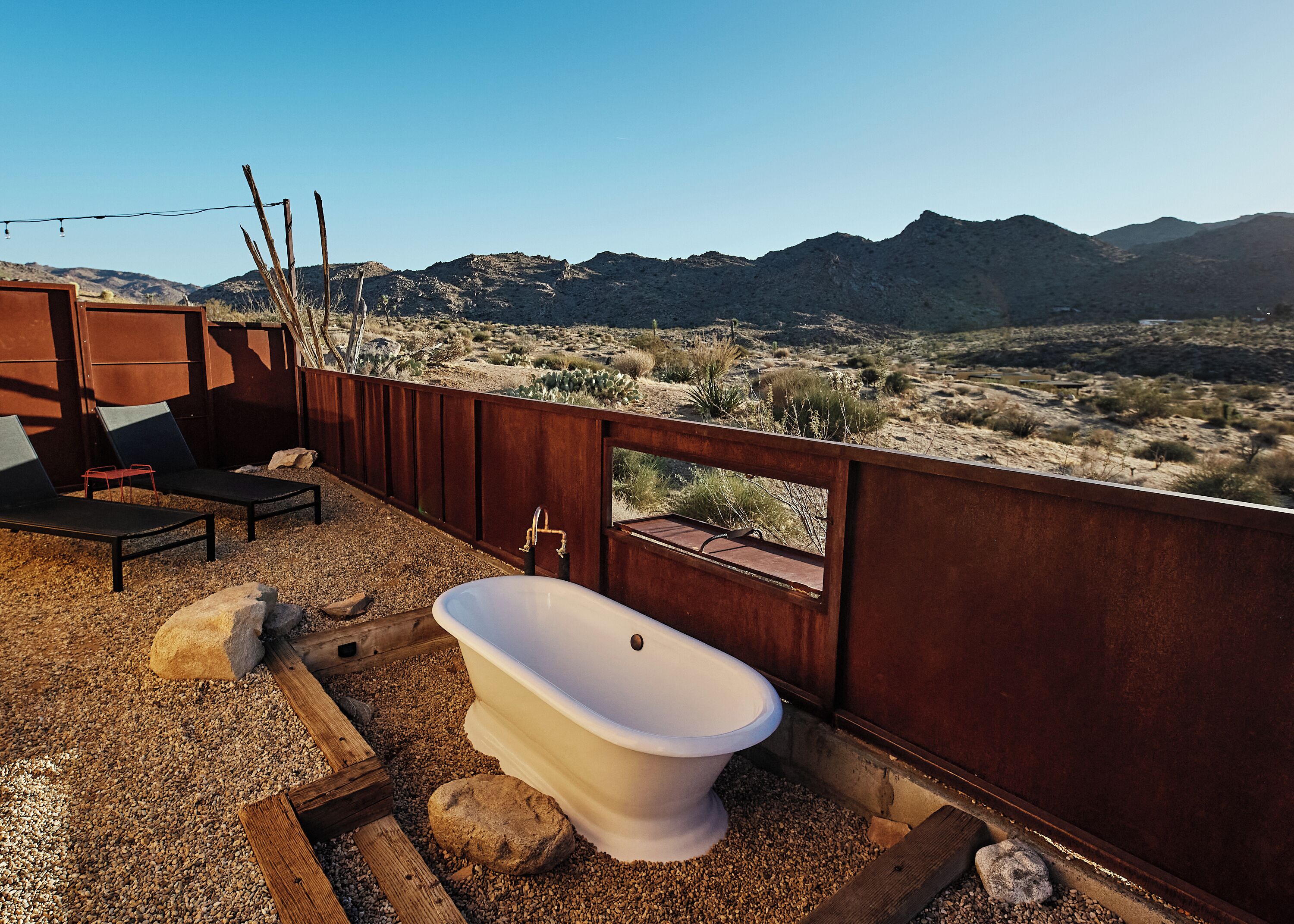 A rustic outdoor bathroom with a white toilet, two lounge chairs, and a view of a desert landscape with mountains in the background.