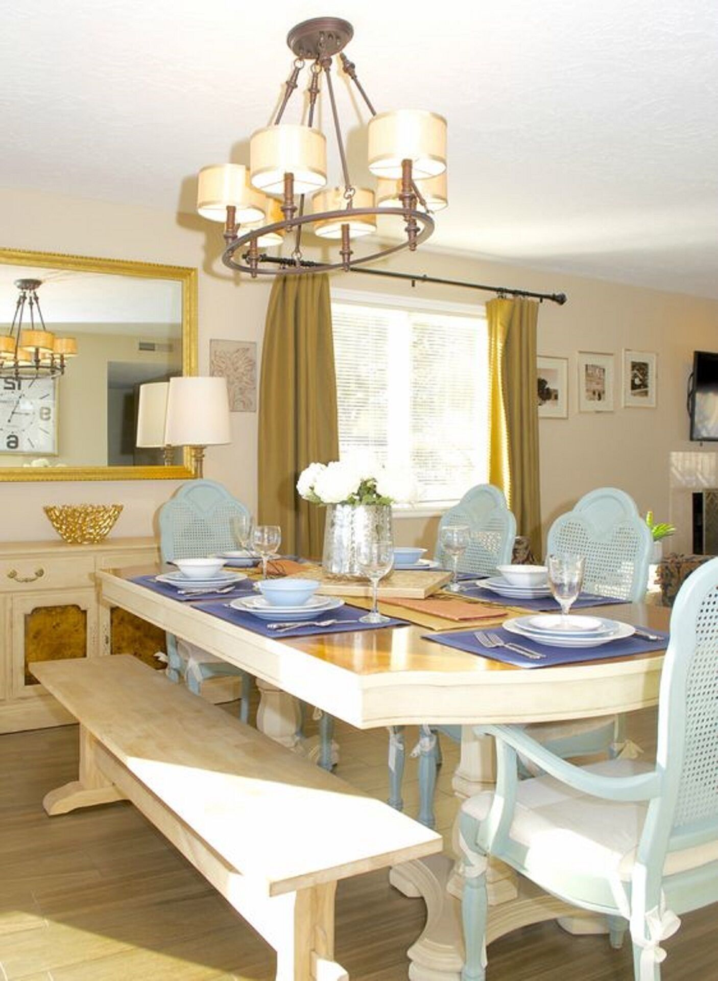 A dining room with a wooden table set for four, blue chairs, a chandelier, a mirror, and a window with curtains.