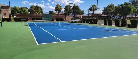 Una cancha de tenis con red, rodeada por una valla y árboles al fondo.