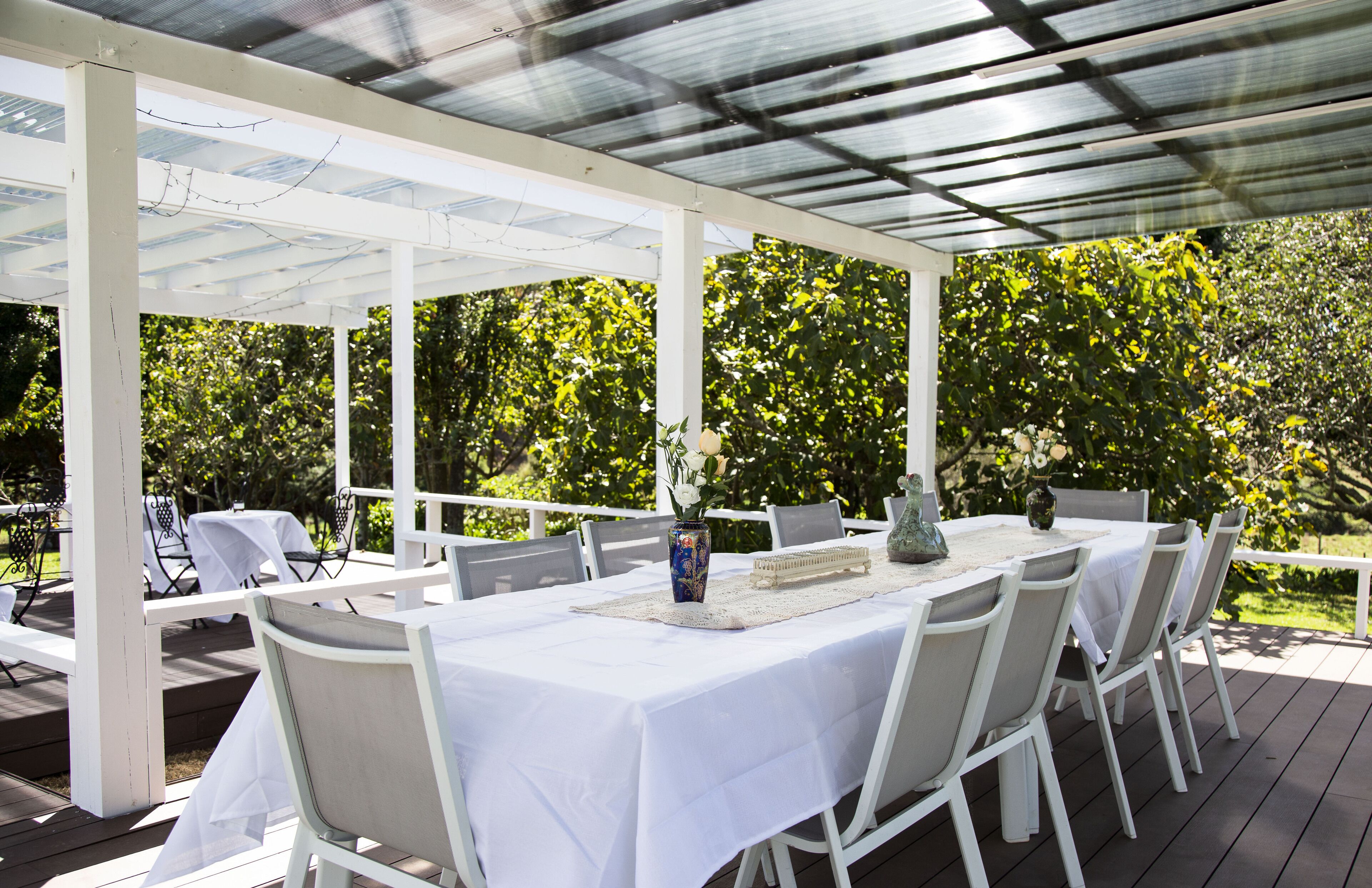 An outdoor patio with white tables and chairs, covered by a glass roof, surrounded by lush greenery.