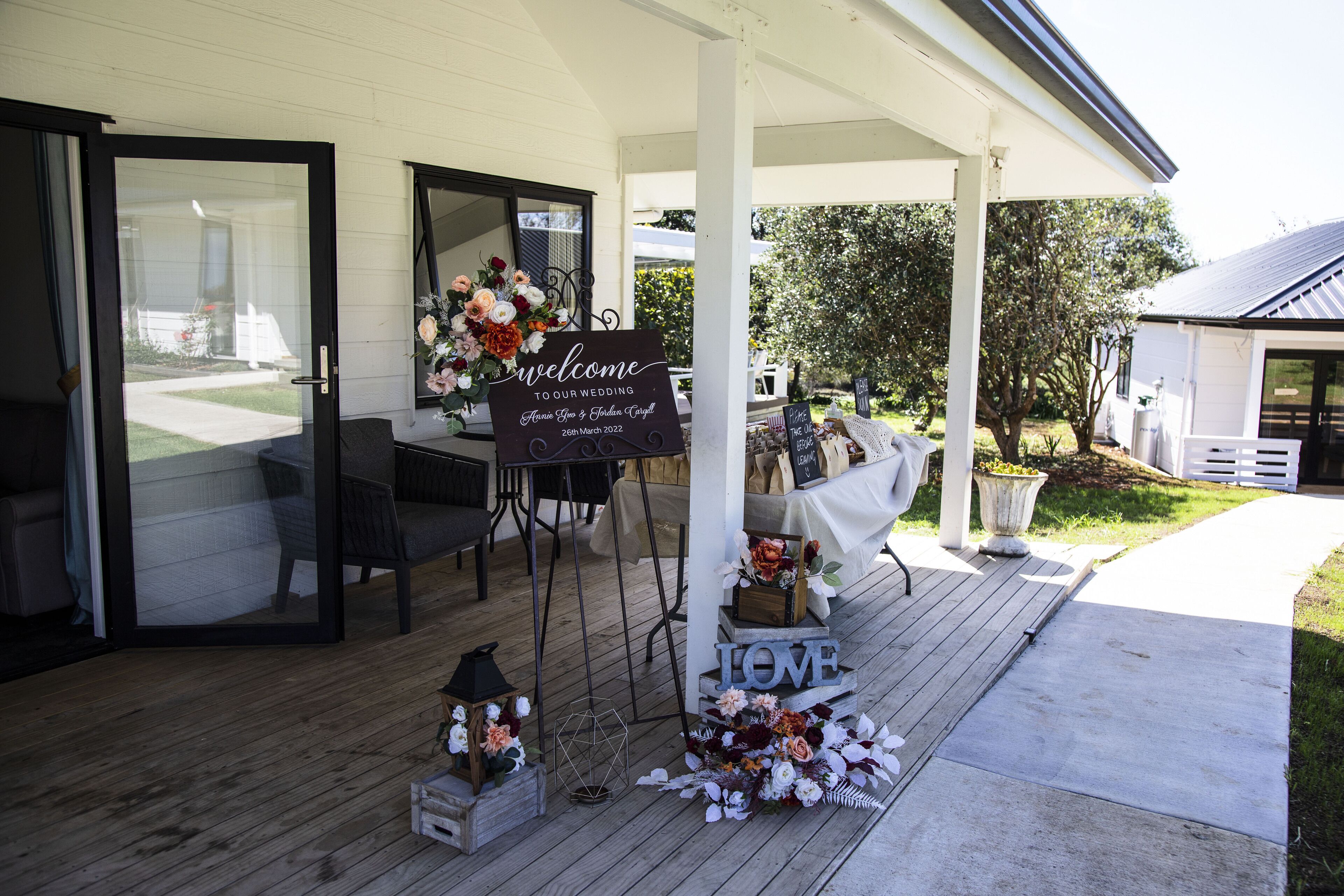 A wedding reception setup with a welcome sign, floral arrangements, and a 'LOVE' decoration on a wooden deck.