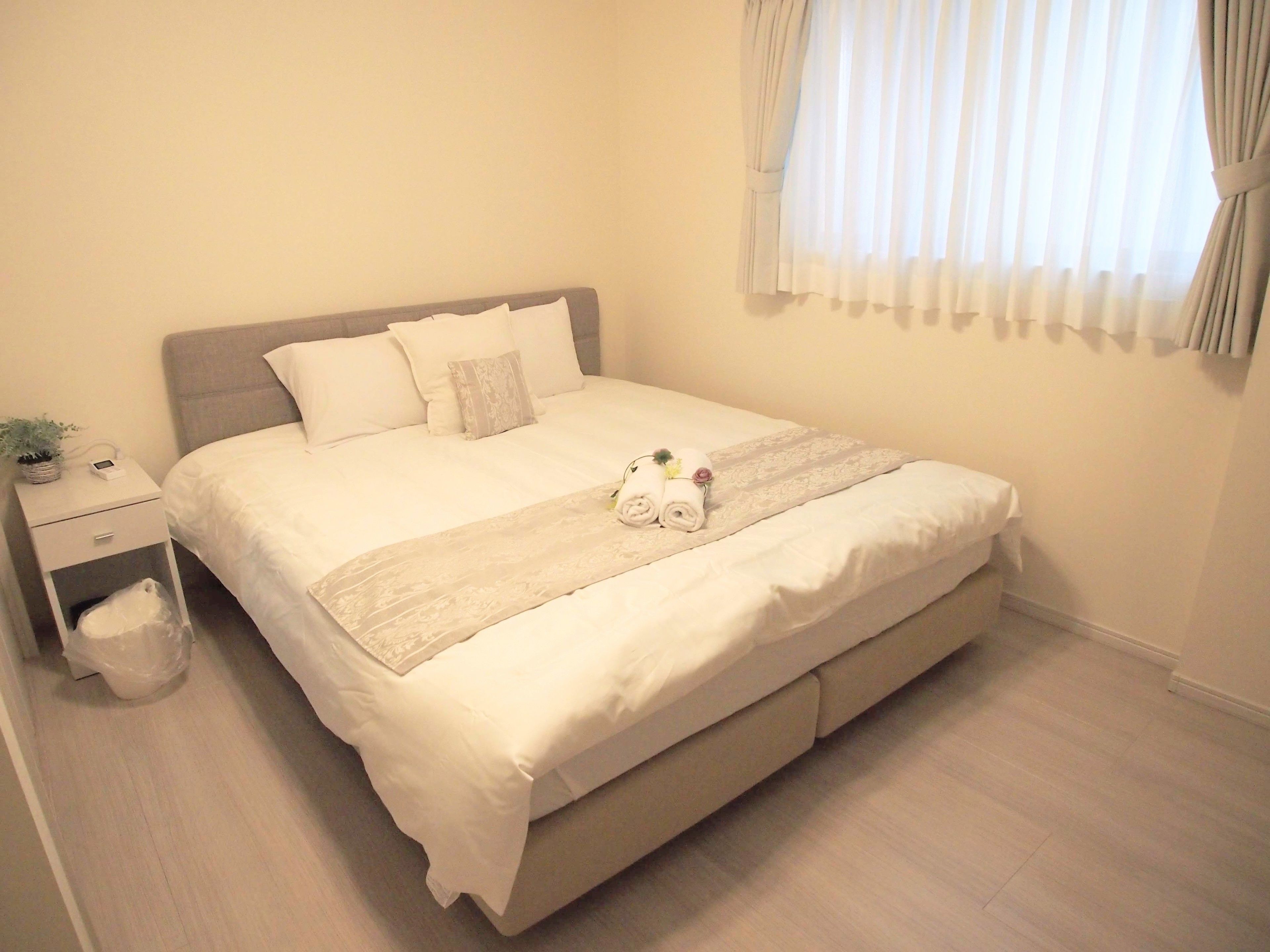 A neatly made bed with a white comforter and pillows, a bedside table with a plant and a lamp, and a window with sheer curtains.