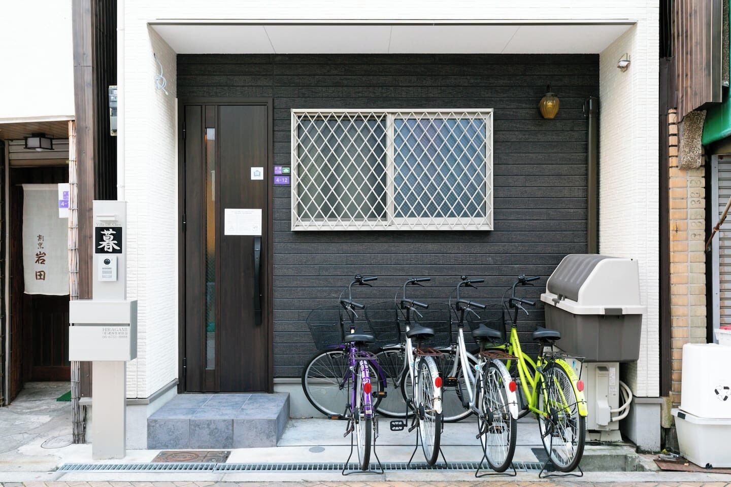 A building entrance with a dark door, a window with a grid pattern, and several bicycles parked outside.