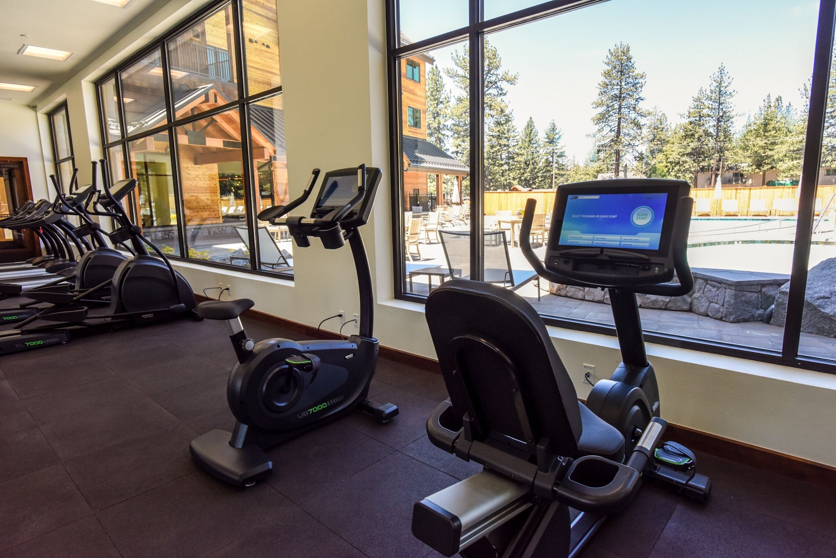A gym with treadmills and an elliptical machine, large windows with a view of outdoor seating and trees.