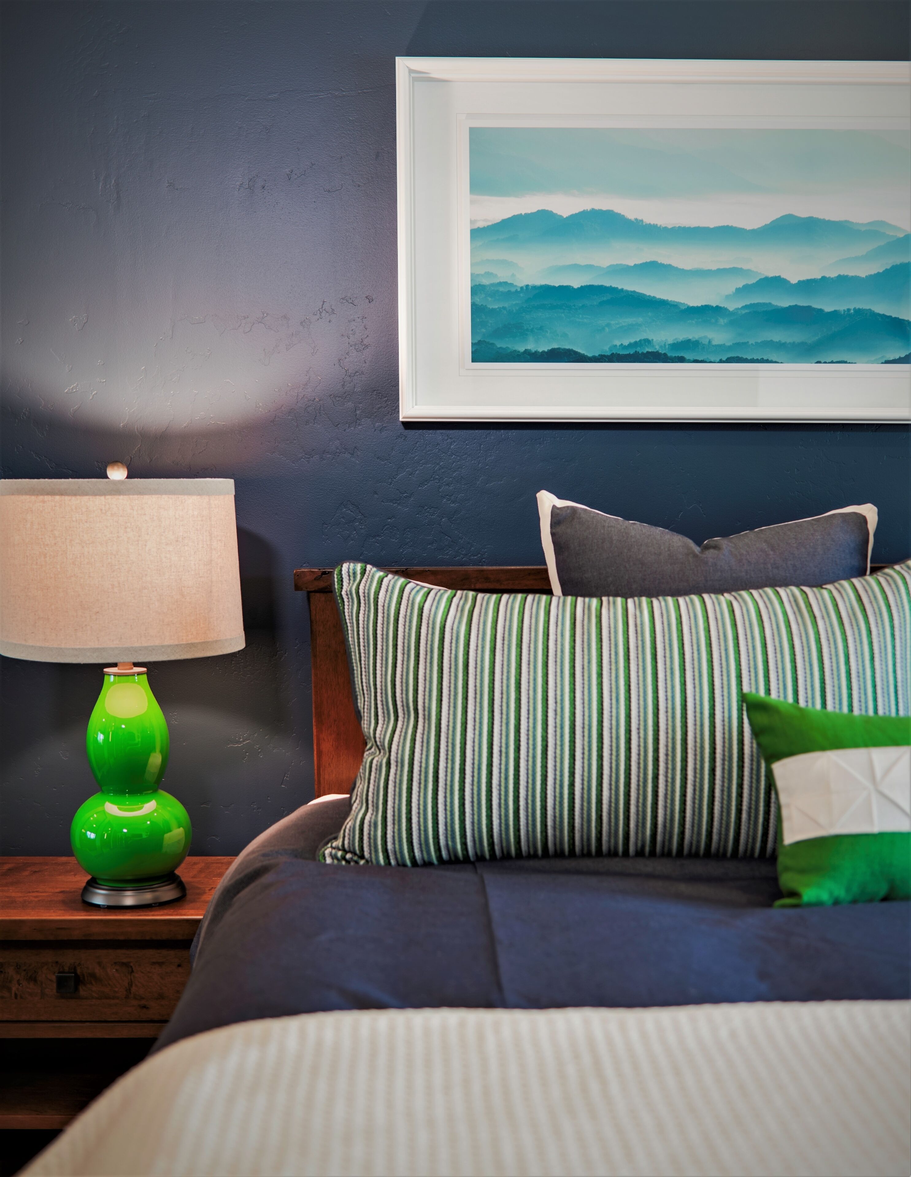 Een netjes opgemaakt bed met gestreepte kussens, een groene lamp op een houten nachtkastje, en een lijst met een landschapsfoto aan de muur.