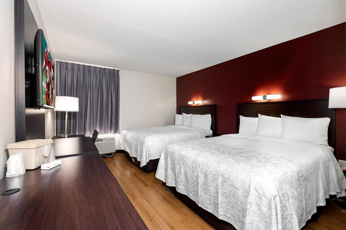 A hotel room with two beds, a desk with a lamp, a TV, and a large window with curtains.
