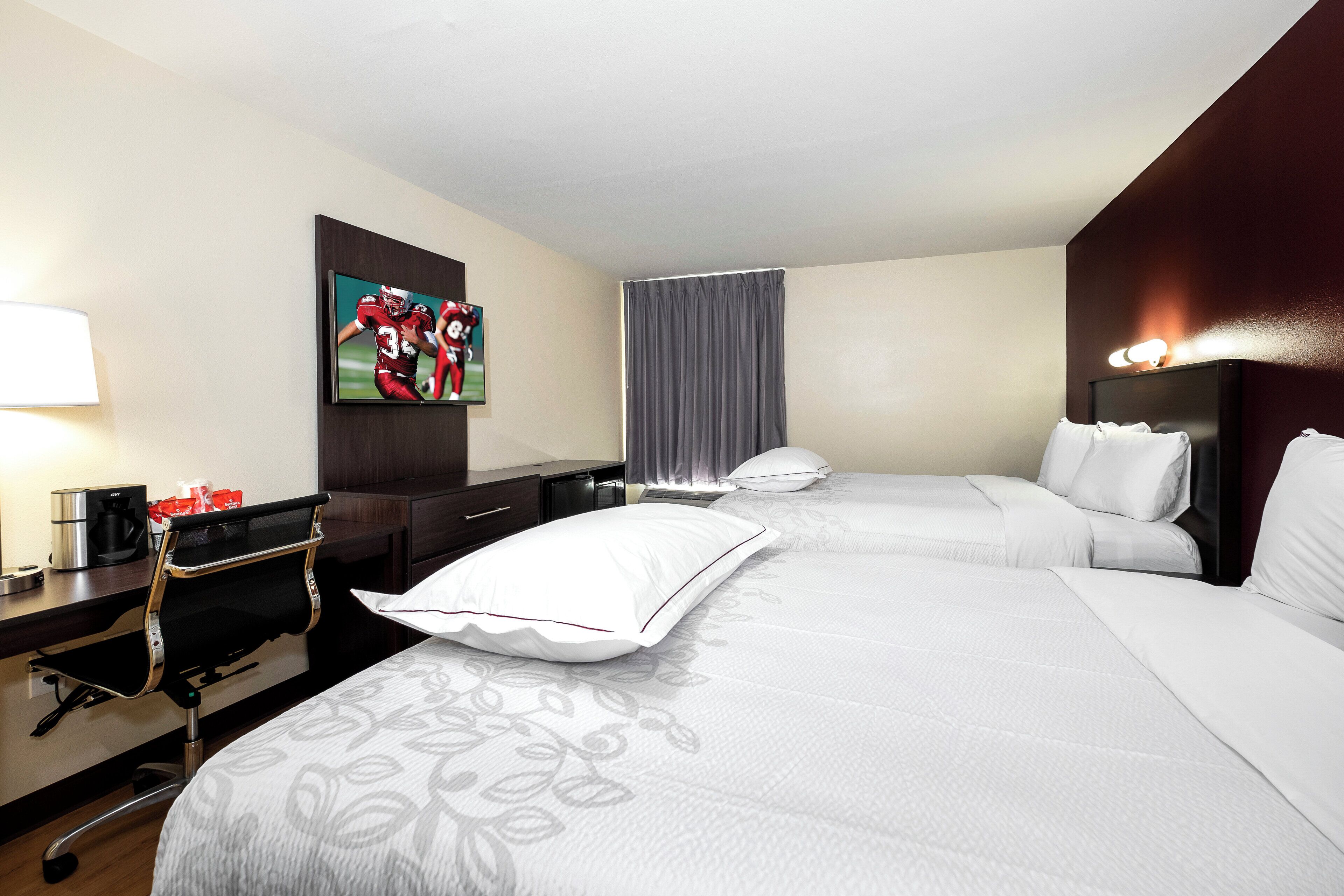 Una habitación de hotel con cama, escritorio con cafetera, televisor en la pared y ventana con cortinas.