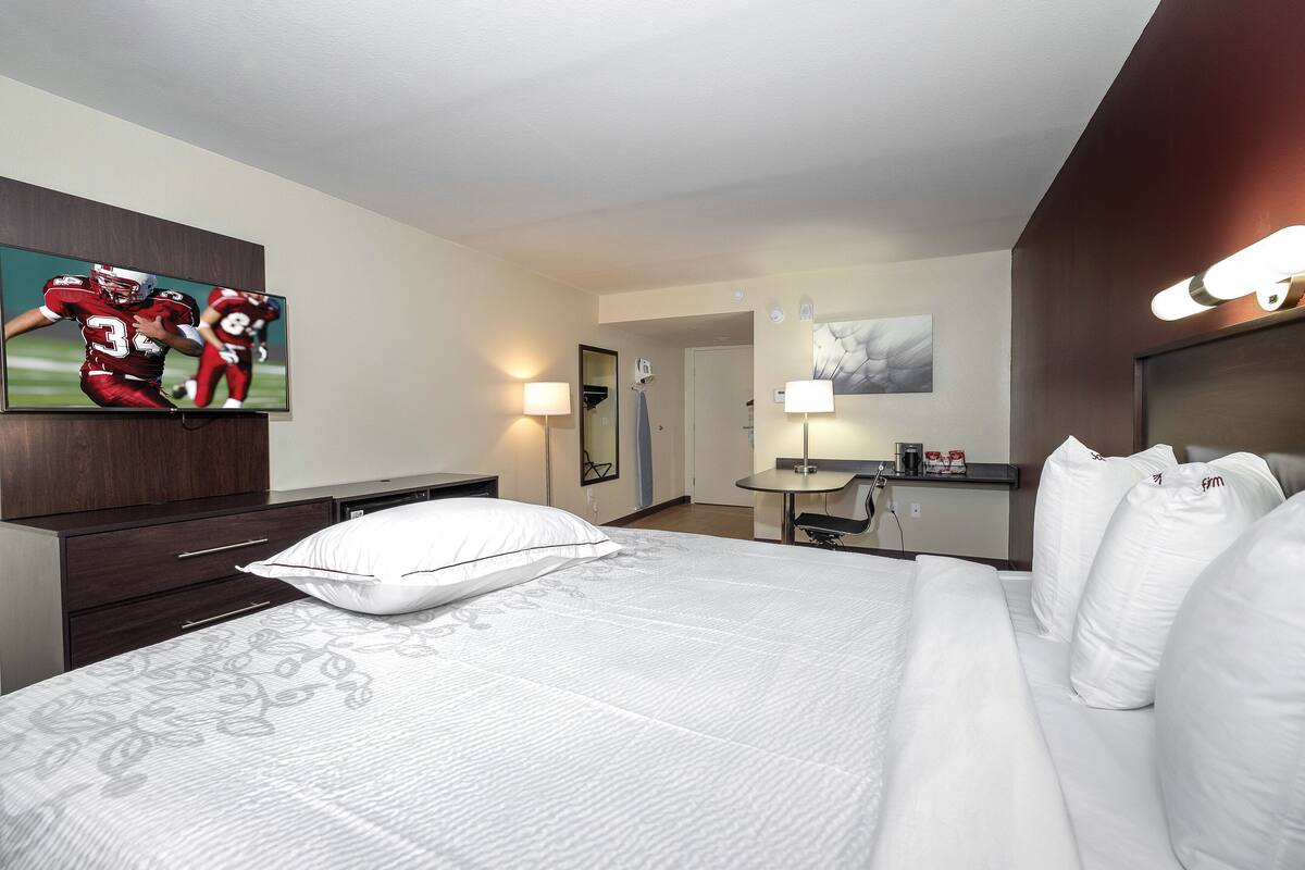 A hotel room with a large bed, a TV on the wall, a desk with a lamp, and a mirror.
