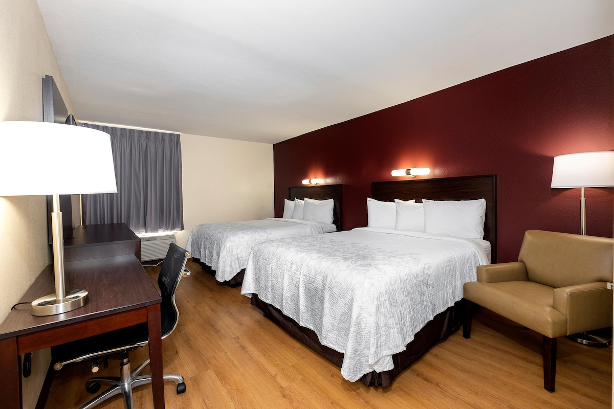 A hotel room with two beds, a desk, a chair, and a lamp.