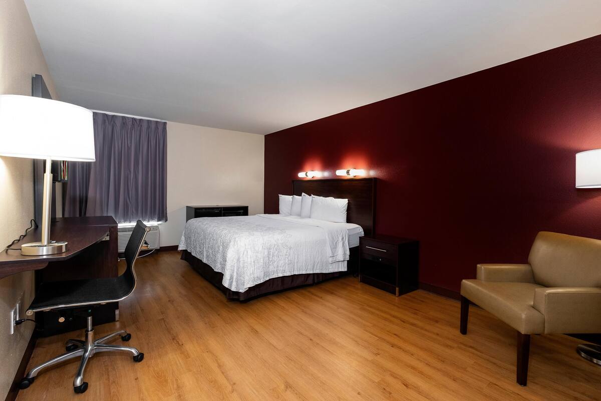 A hotel room with a bed, desk, chair, and a red accent wall.