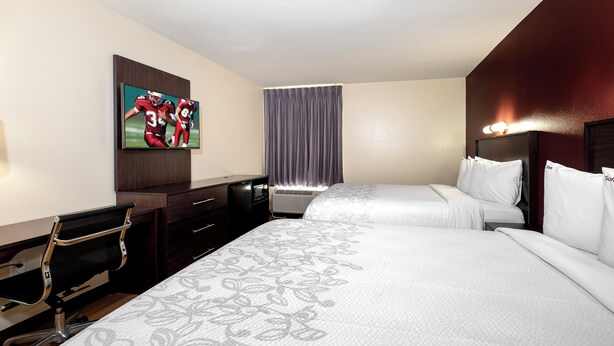 A hotel room with two beds, a desk with a chair, a flat-screen TV, a window with curtains, and a wall-mounted light fixture. - Red Roof Inn PLUS+ Tuscaloosa - University (Tuscaloosa)