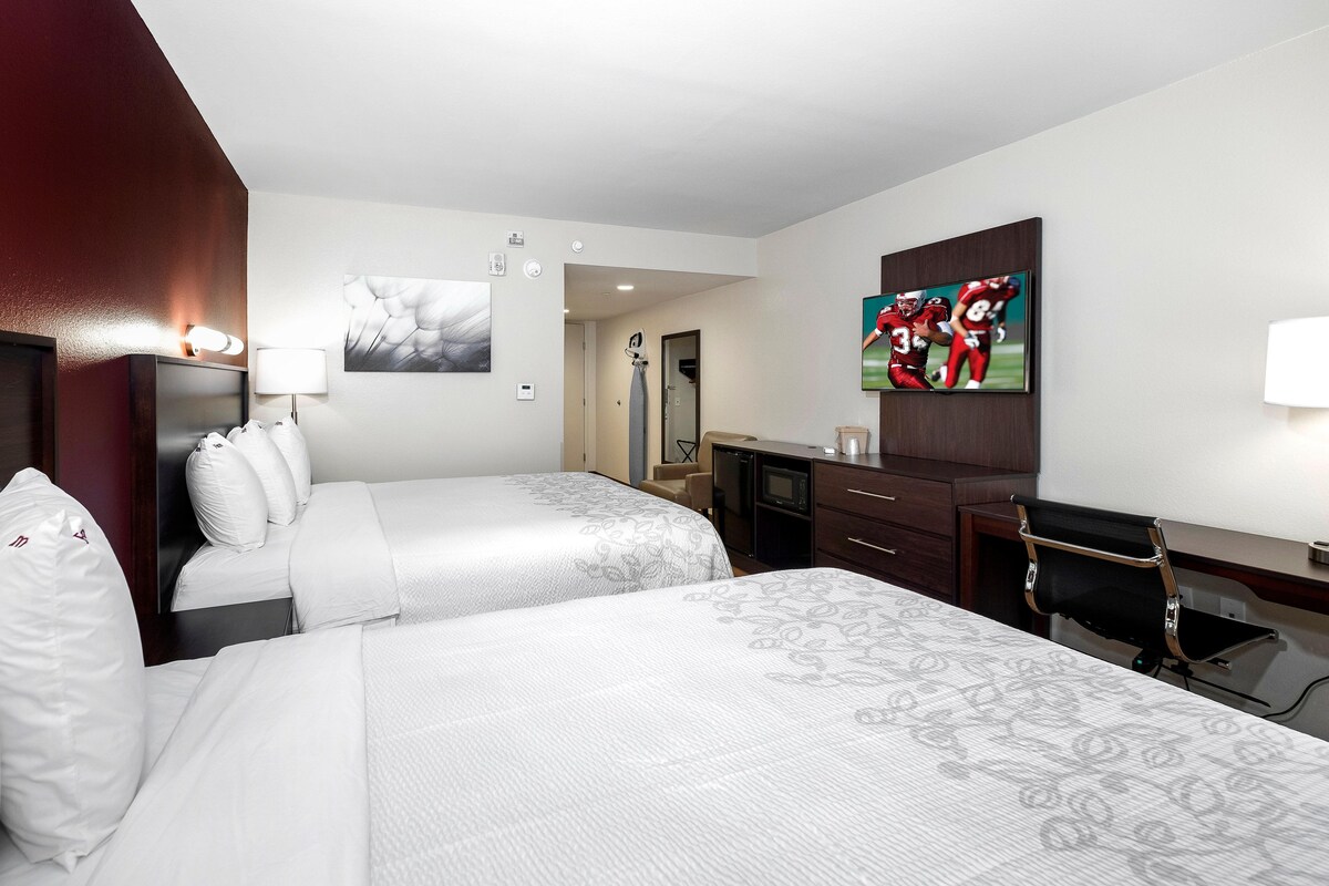 A hotel room with two beds, a desk, a chair, a TV, and a painting on the wall.