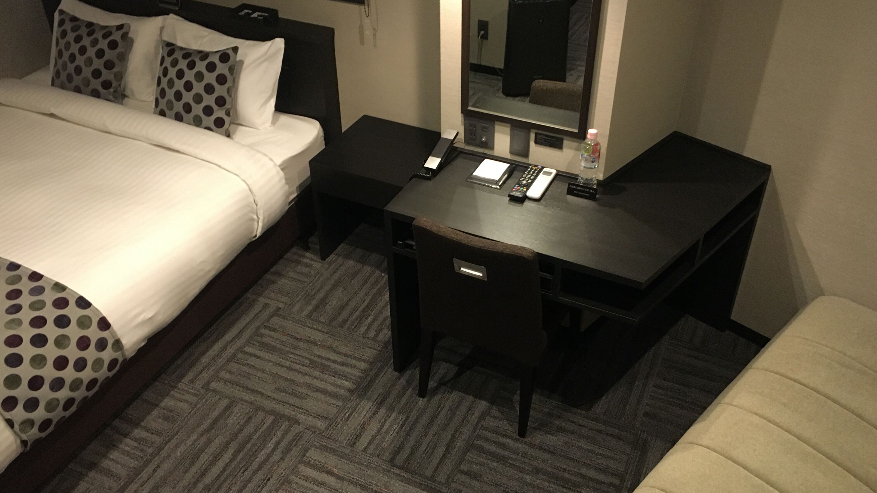 Ein Hotelzimmer mit einem Bett, einem Schreibtisch, einem Stuhl, einem Spiegel und einem kleinen Tisch mit einer Flasche.