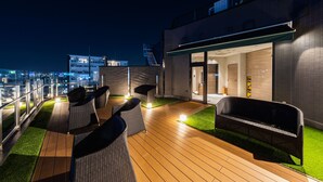 A rooftop terrace with wicker furniture, a wooden deck, and a glass balustrade overlooking a cityscape at night. - Astil Hotel Juso Precious (Osaka)