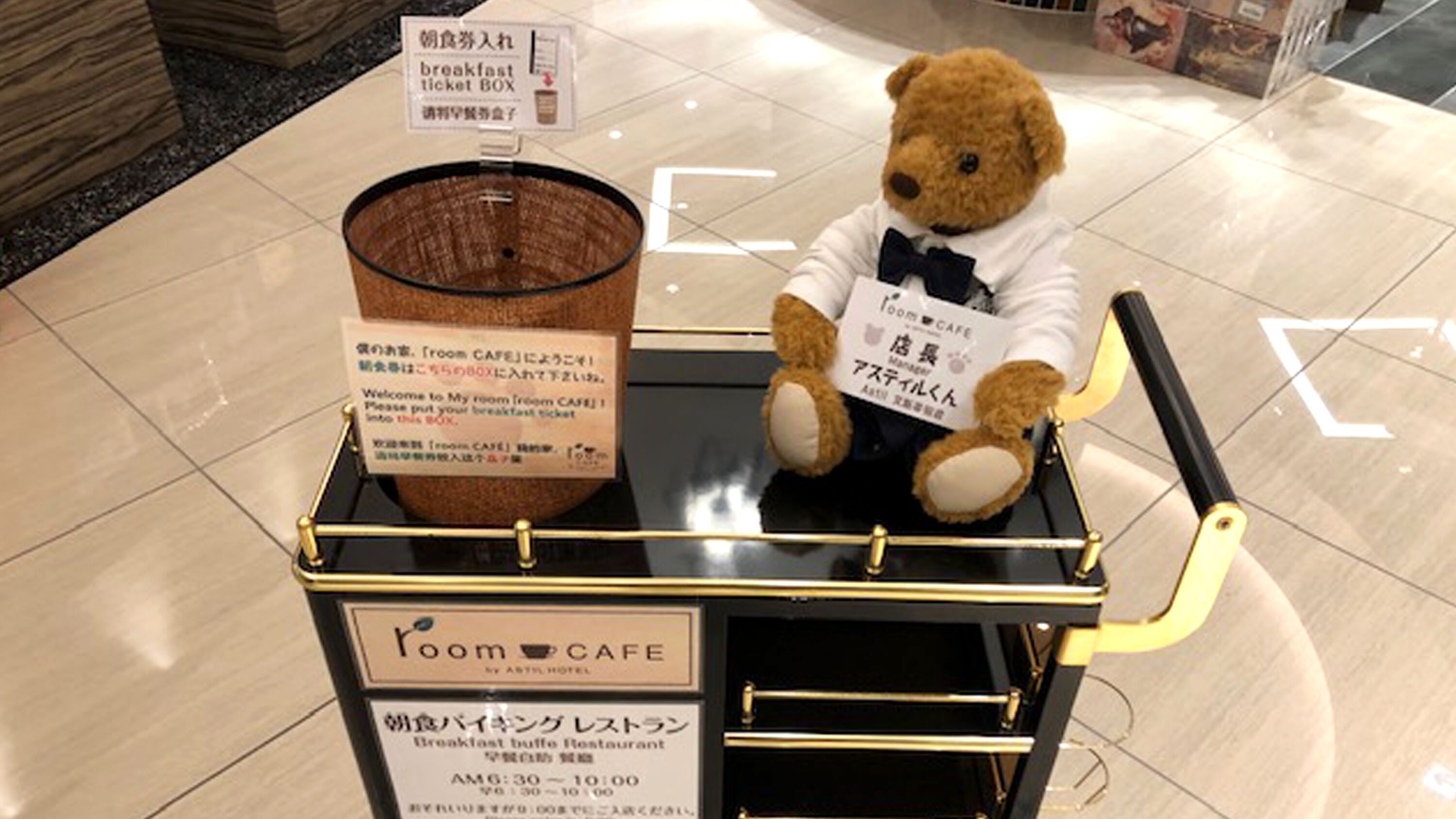 A teddy bear in a suit sitting on a cart with a sign that reads "breakfast ticket BOX" and "Room Cafe".