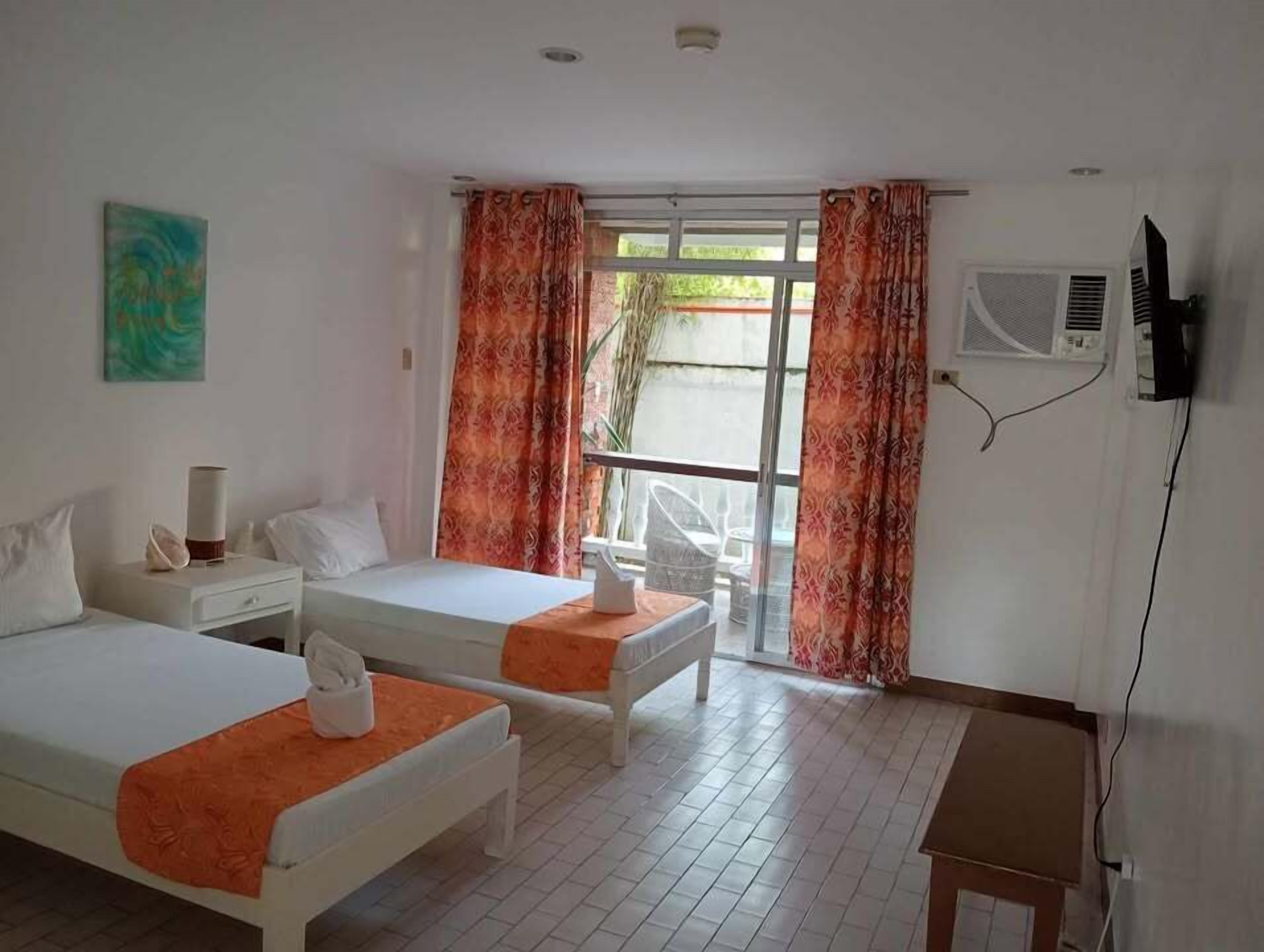 A hotel room with two beds, a small table, a chair, a TV, and a window with curtains.