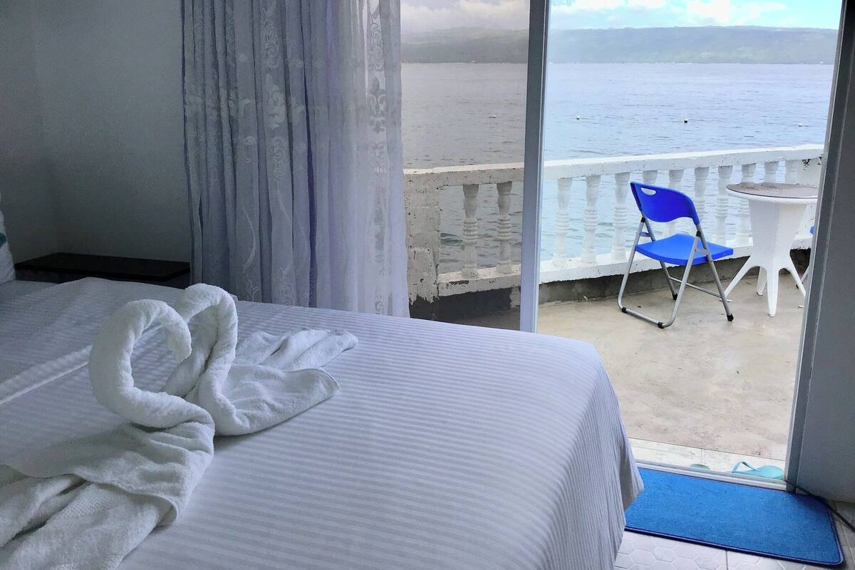 A hotel room with a bed, a white towel, a balcony with a blue chair, and a view of the sea.