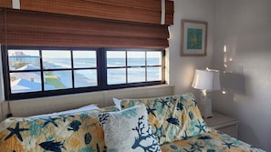 A hotel room with a bed, a window with a view of the sea, a lamp, and a framed picture on the wall.
