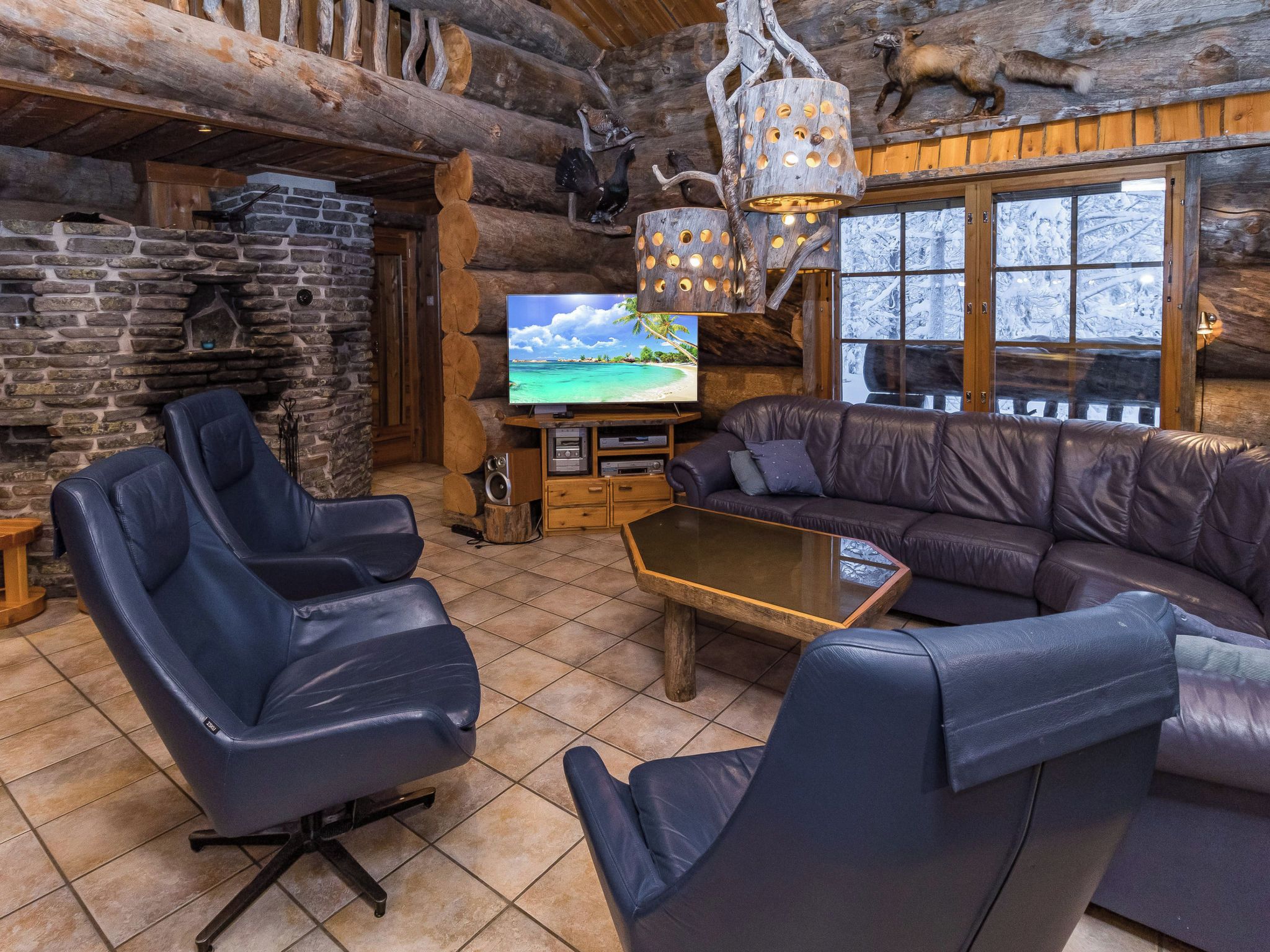 A log cabin interior with a stone fireplace, leather seating, a television, and a large window.