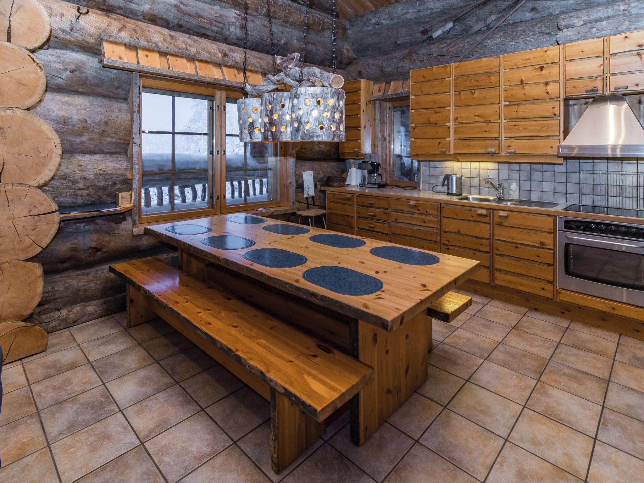 L’intérieur d’un chalet rustique est aménagé avec une table à manger en bois, des bancs, une cuisine dotée de meubles en bois et une grande fenêtre donnant sur un paysage enneigé.