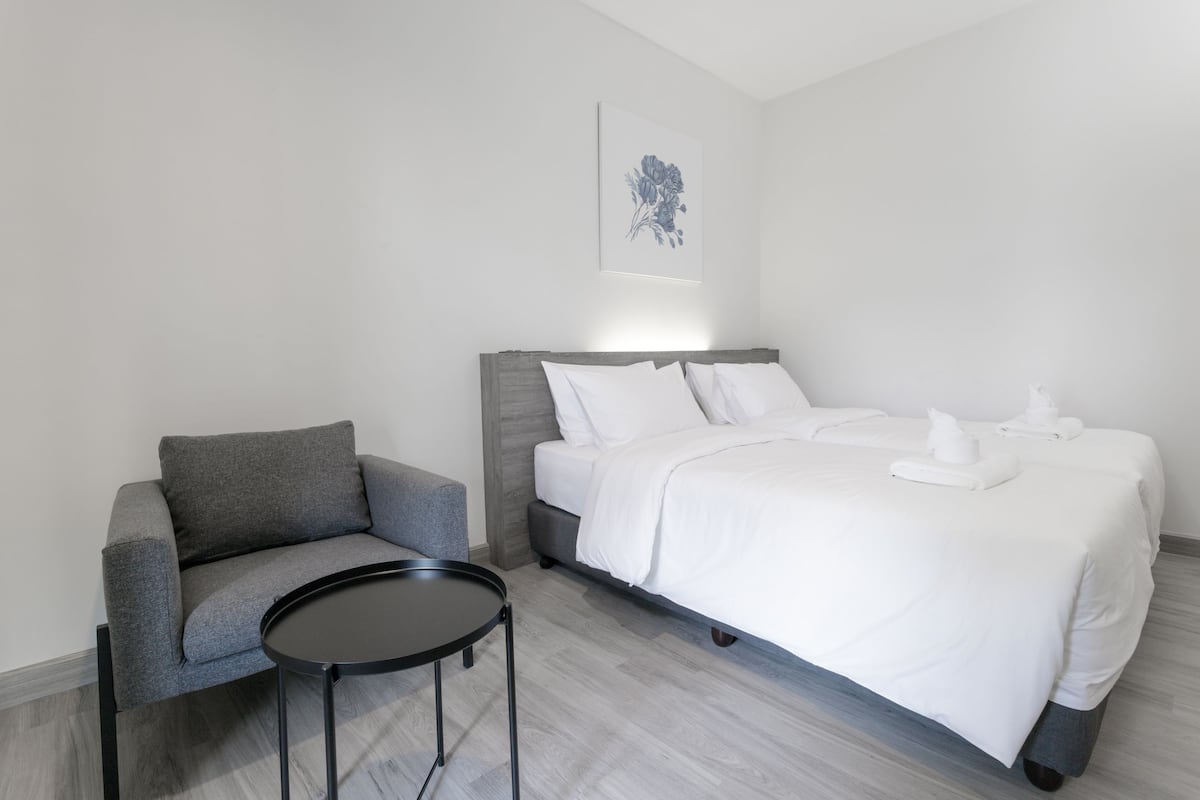 A modern hotel room with a grey sofa, a small round table, a large bed with white linens, and a wall-mounted artwork.