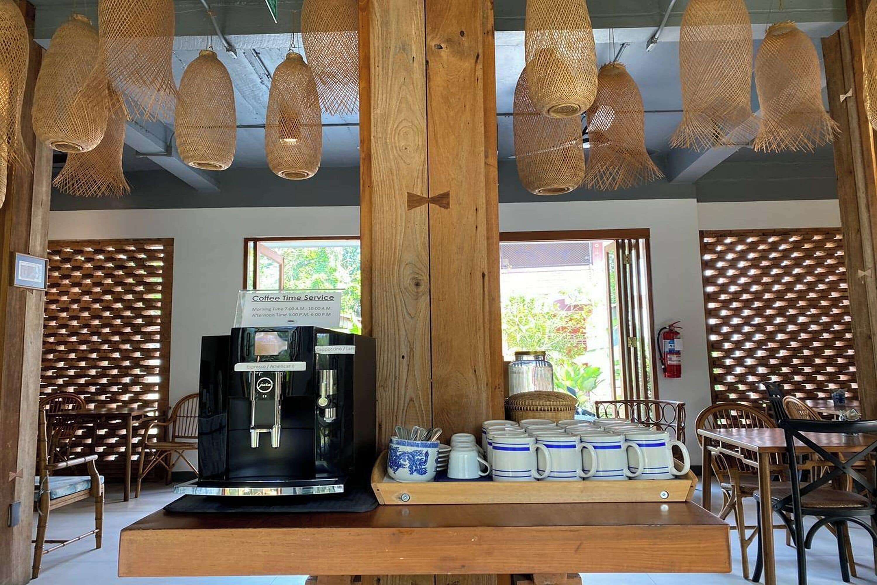 A coffee shop interior with a coffee machine, wooden tables, wicker hanging lights, and a view of greenery outside.