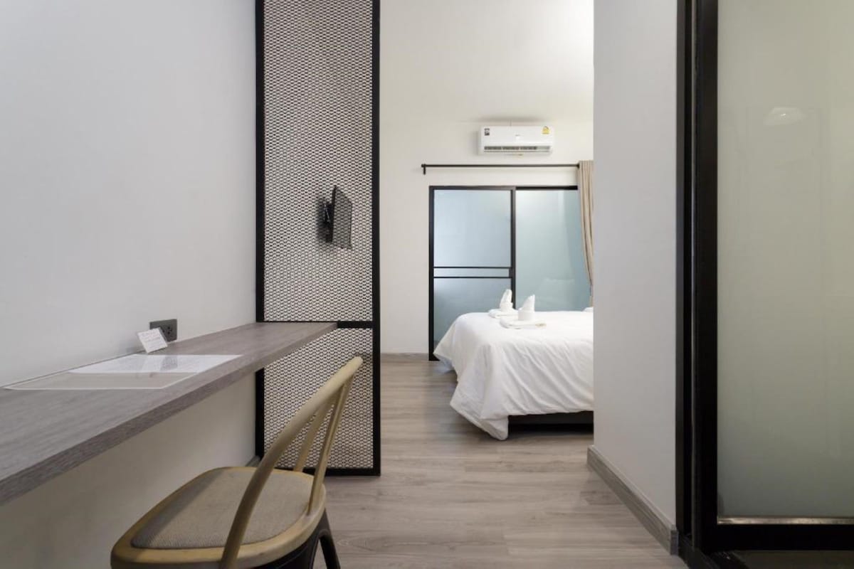 Una habitación de hotel moderna con un escritorio de madera, una silla, una cama con ropa blanca y un ventanal grande.