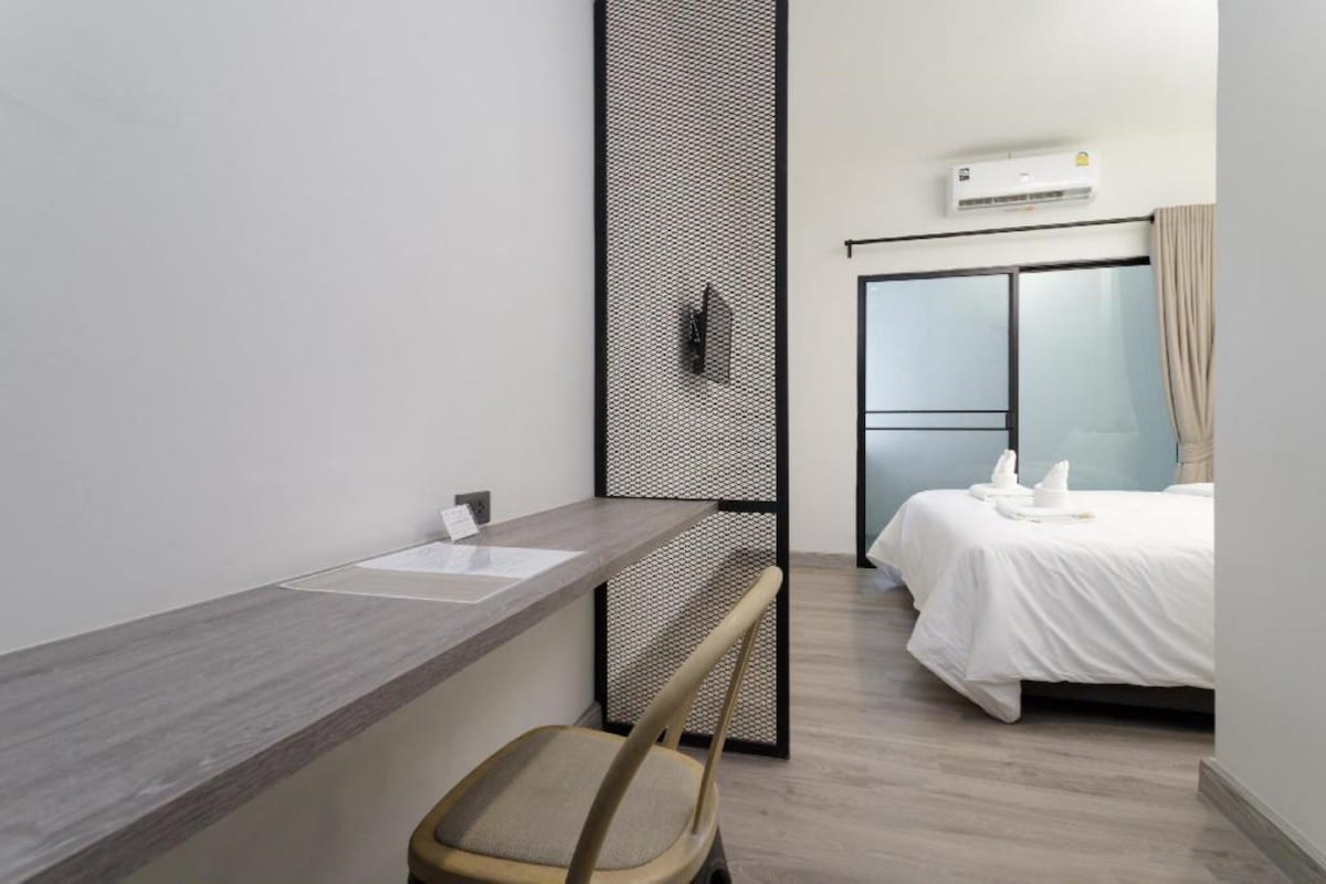A modern hotel room with a wooden desk, a chair, a bed with white linens, a wardrobe, and an air conditioning unit.