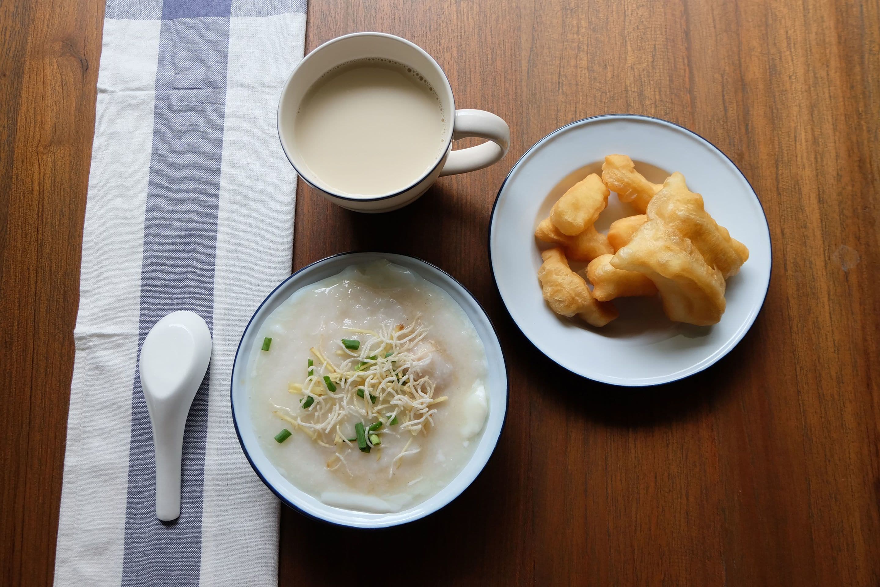 A wooden table with a white plate of fried dough, a bowl of soup with green onions, and a white mug.