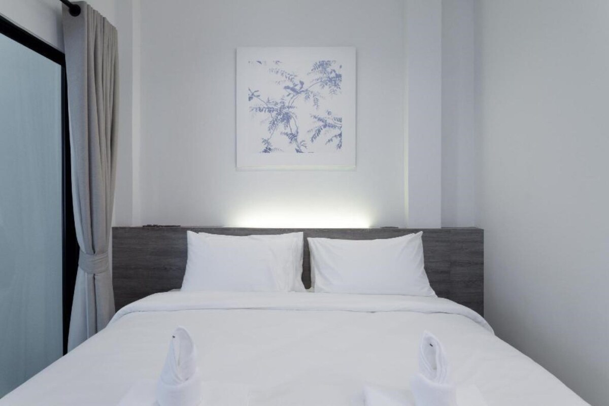 A neatly made bed with white linens and pillows, a headboard with a dark wood finish, and a wall-mounted artwork depicting abstract branches.