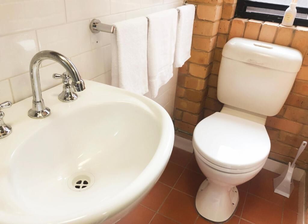 Ein Badezimmer mit weißem Waschbecken, einem Chrom-Wasserhahn, einer Toilette und einem Fenster zur Ziegelwand.