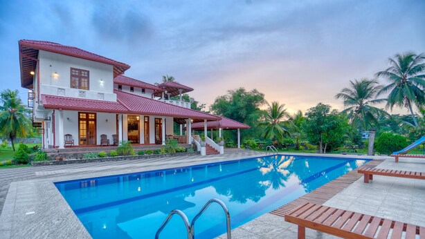 A large swimming pool with a wooden deck and lounge chairs, surrounded by lush greenery and a two-story house with a red roof. - Om Residence (Beruwela)