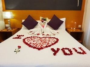 Habitación de hotel con una cama decorada con pétalos de rosa formando un corazón y la frase 'I love you' escrita.