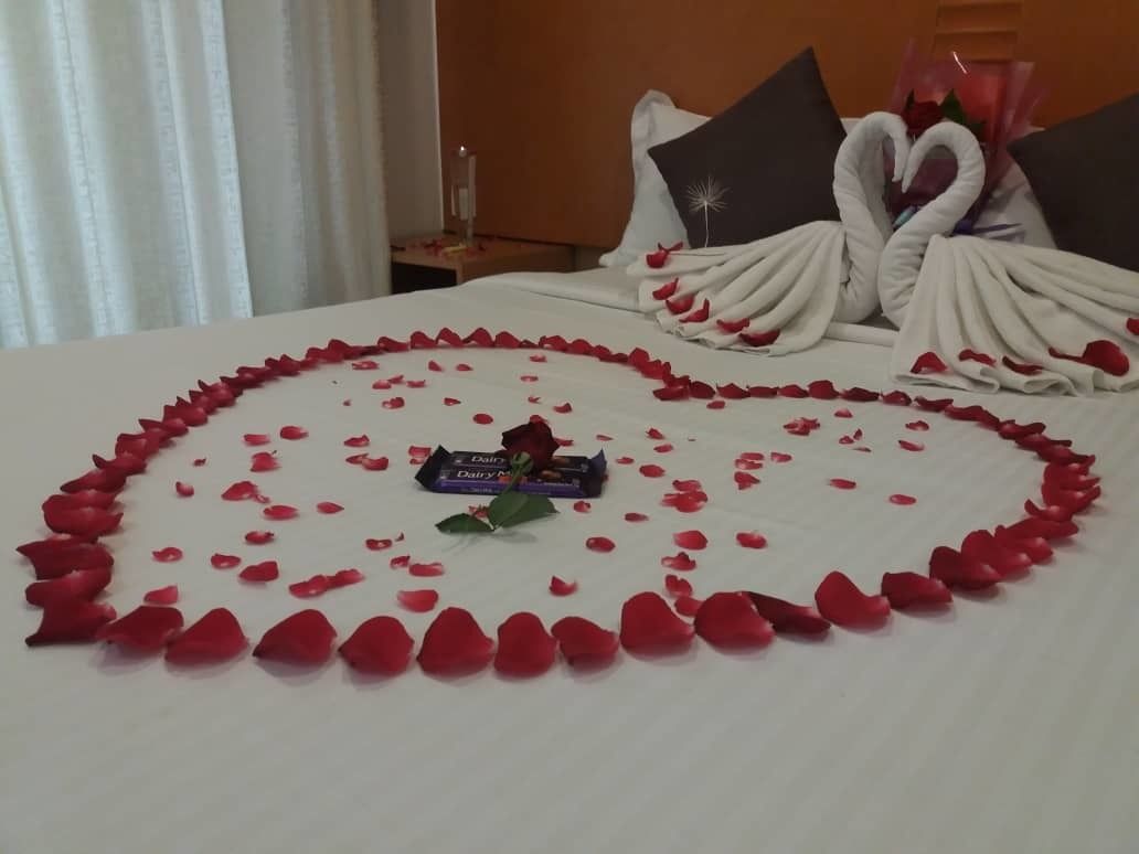 A bed with rose petals arranged in a heart shape, a chocolate bar, and a bouquet of roses.