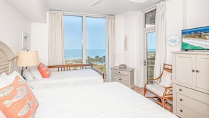 A hotel room with two beds, a TV, a desk, and a view of the ocean.
