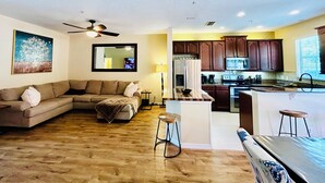 Ein Wohnzimmer mit einer Couch, einem Couchtisch und einem Fernseher. Es gibt eine Küche mit Kühlschrank, Herd und Schränken.