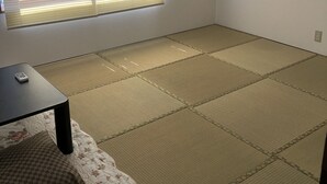 A room with tatami flooring, a small table with a remote control, a window with blinds, and an air conditioning unit. - Yoko House (Uruma)
