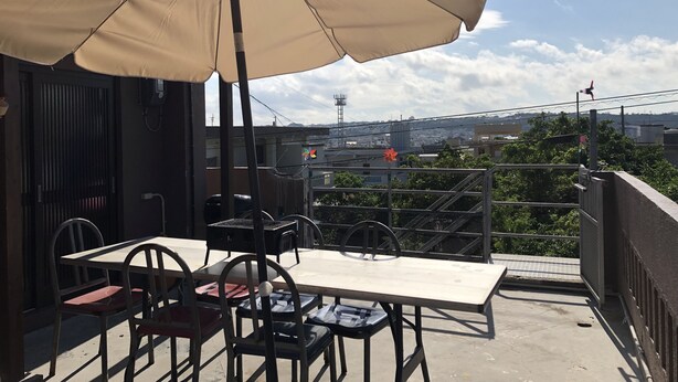 A balcony with a table and chairs, a large umbrella, and a view of the cityscape. - Yoko House (Uruma)