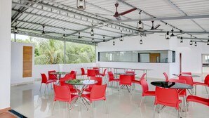 Um espaço amplo e interno, com cadeiras e mesas vermelhas, janelas grandes e um teto alto com ventiladores e luminárias.