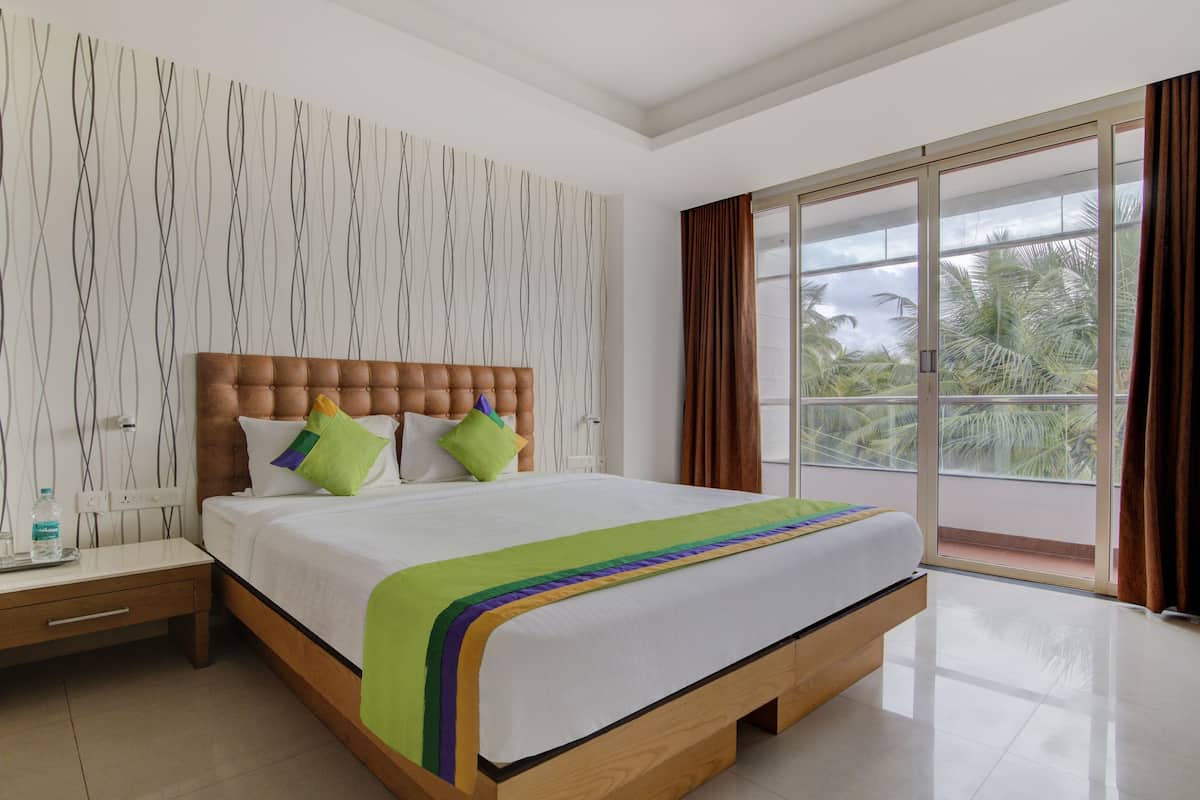 A hotel room with a large bed, a nightstand, a window with curtains, and a view of palm trees.