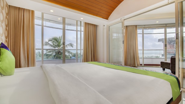 Quarto de hotel moderno com uma cama grande, janelas amplas e vista para palmeiras.