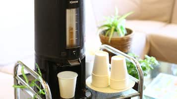 Uma cafeteira de café preto sobre um carrinho, com uma mesa de vidro, chávenas brancas e uma planta em vaso.