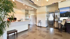 A hotel reception area with a reception desk, a clock, a plant, a bench, and a glass elevator. - Chino Sky View Hotel (Chino)