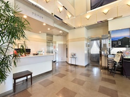 Zona de recepción de un hotel con mostrador de recepción, un reloj, una planta, un banco y un ascensor de vidrio.